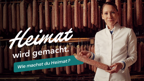 Heimatmacherin Katharina Koch im Vordergrund wie sie stolz eine Wurst in den Händen hält. Im Hintergrund sehen wir viele hängende Würste zum trocknen. Zudem der Slogan "Heimat wird gemacht. Wie machst du Heimat?"