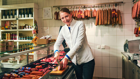 Das Bild zeigt Katharina Koch in ihrer Landfleischerei wie sie gerade eine Wurst schneidet. Im Hintergrund sieht man eine mit Fleisch und Wurstwaren gefüllte Theke sowie ein Regal mit Verkaufsprodukten und weitere Würste, die an der Wand hängen.